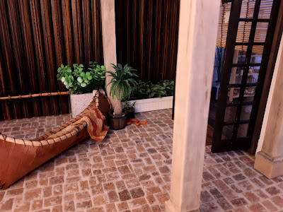 One twelfth scale modern courtyard with paved flooring, a bamboo fence, a selection of pot plants and a canoe with a retro towel draped over it. A snake is slithering out from between two of the pot plants towards the open french doors