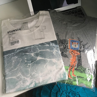 Two t shirts in clear packaging, one has a picture of the sea on it and the other a drawing of a human body with a cat's head, and a box on its head