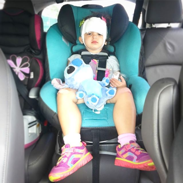 Toddler girl with bandages wrapped around her head frowns while sitting in her carseat