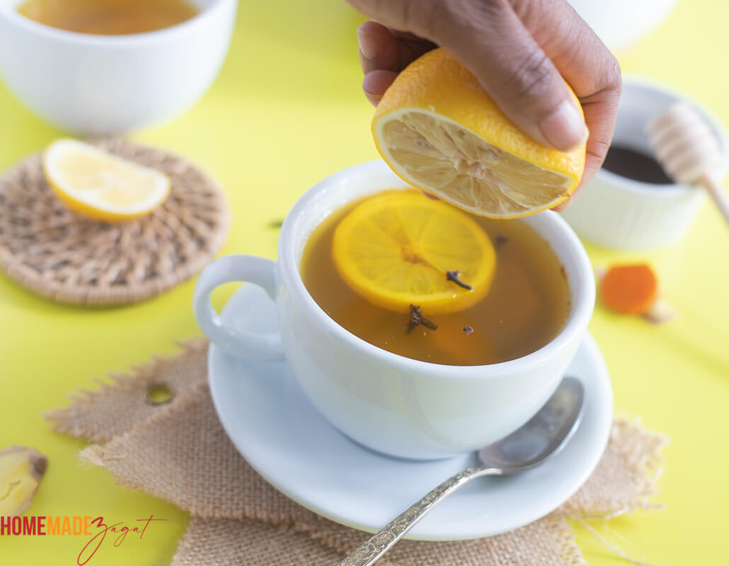 A hand squeezing some fresh lemon juice into white teacup of turmeric ginger lemon tea with slices of lemon