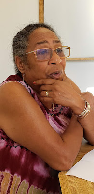 ID: the Rev. Dr. Pero in a purple and pink sleeveless dress, with her slightly graying hair pulled back into a ponytail. She's sitting in front of a white background at a woodgrain colored table with her elbow on the table and her hand on her chin. Her other hand reaches around her elbow.
