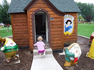 a toddler with a purple skirt and short curly red hair walks into a wooden cabin with Snow White's face on it and statues of dwarves outside it