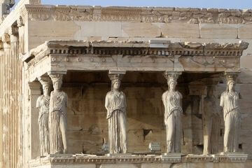 Fotografia das Cariátides do templo do Erecteion: Trata-se de uma construção feita de pedra, com uma marquise sustentada por várias cariátides, que são esculturas de mulheres sustentando a marquise ao invés de pilastas. A cor da construção é bege, tendo variações de tonalidade. Acima, ao fundo se vê o pedaço do céu, um tom azul claro.