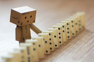 A robot made of amazon boxes stands next to a line of domino tiles that are standing up. The robot has a hand extended to one tile as if to tip it and cause the others to fall.