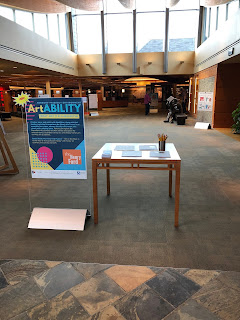 view of open air lobby with sky lights and brown tile and carpet floor. There is a poster stand with a blue poster inside reading ArtABILITY next to a wood table with handouts and pencil holder. Ranges of shelving to the left and a service desk are in the background.