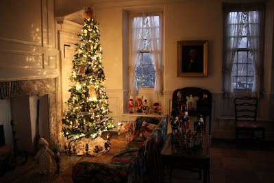 Tall slender Christmas tree with white lights and ornaments stands next to the marble fireplace. A dozen or more nutracker figures of different sizes are placed on window sills and tables around the room.