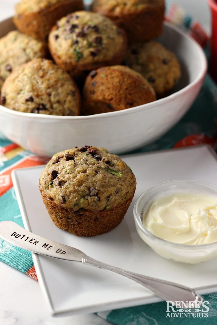 Chocolate Chip Zucchini Muffins by Renee's Kitchen Adventures in a bowl in the background with one muffin in the foreground on a white plate with a spreader for butter resting on the plate