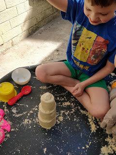 A boy wearing green shorts and a blue top is sitting with his legs crossed and his right hand raised high above a three tiered sand castle