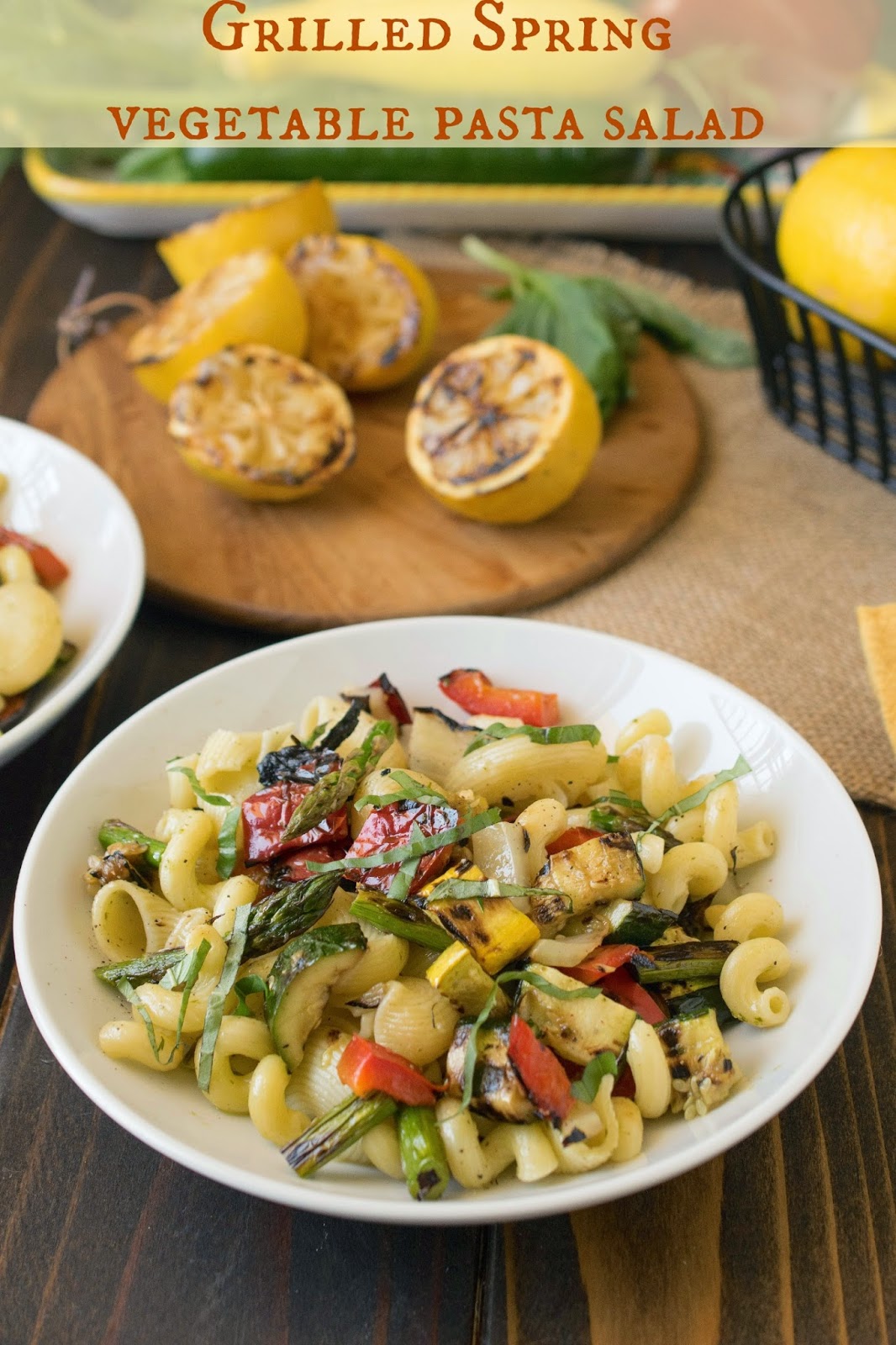 Grilled Spring vegetable pasta salad in a white bowl with grilled lemon halves on a board behind