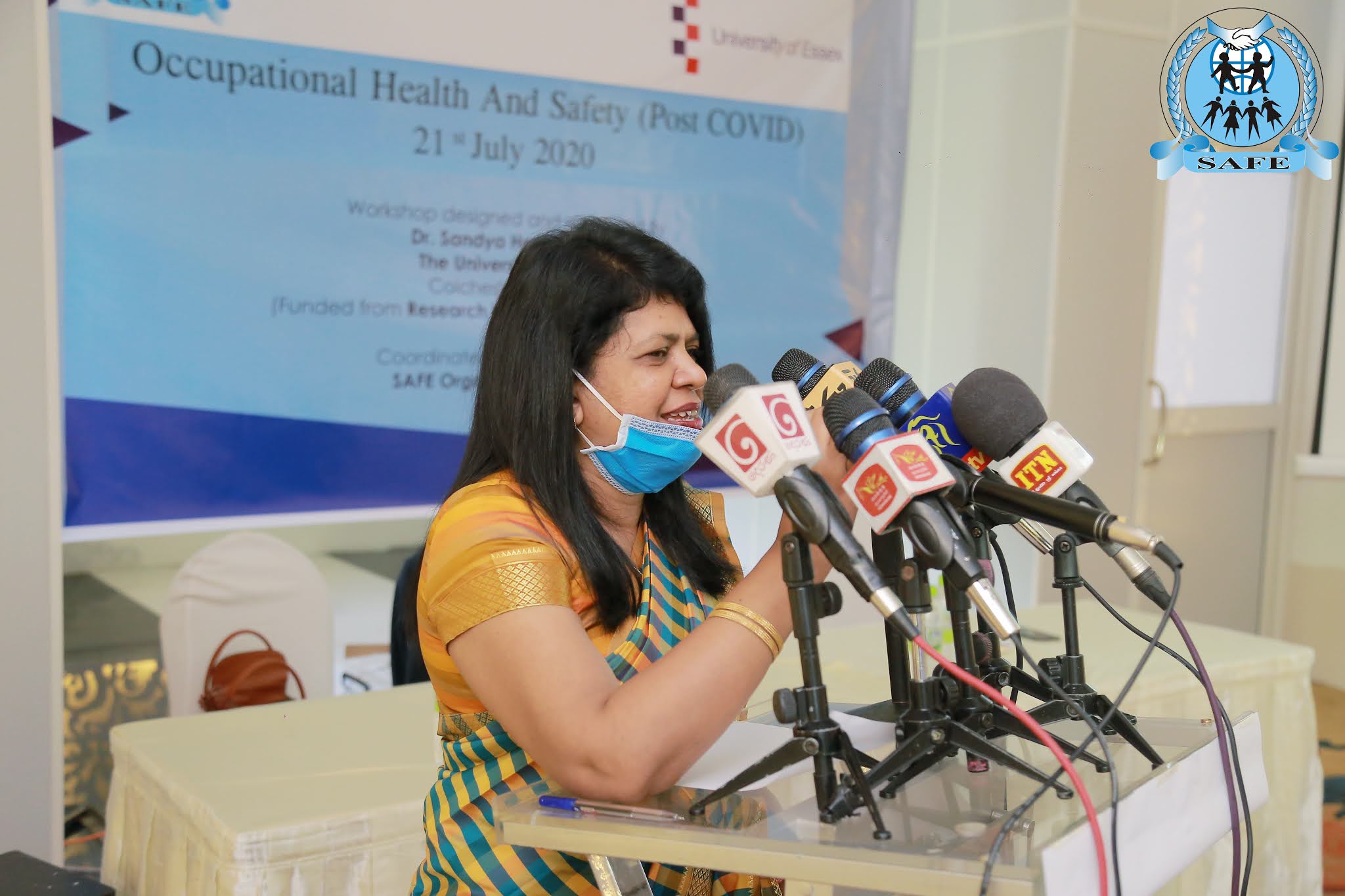  A woman wearing a sari and a face mask is speaking into a microphone with the SAFE and Occupational Health and Safety logos in the background.