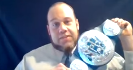 Bearded man in white shirt and blue tie holds a wrestling belt with medallions on it, center medallion has the PHMC logo on it in blue