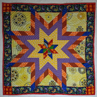 Blue, red, yellow, purple, and orange print fabrics create a modern Lone Star on a pale yellow background printed with large medallions. The navy border has lines of colorful geckos and the corners are posted with and orange on red batik.