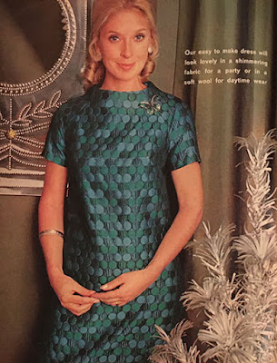 A blonde woman in a green and turquoise brocade shift dress with high round neck