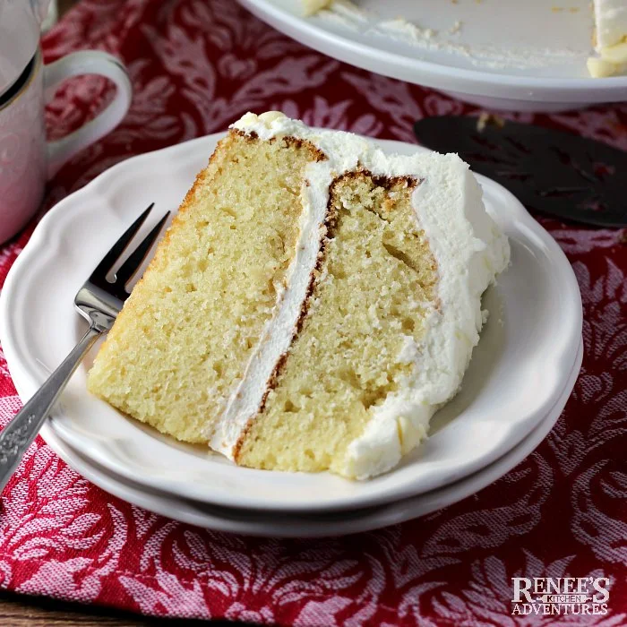 White Chocolate Cake by Renee's Kitchen Adventures slice of cake on white plate with fork, coffee cups in background