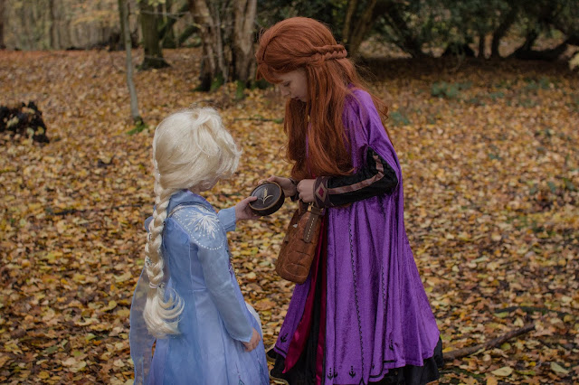 2 girls dressed in Anna and Elsa ShopDisney costumes and wigs from Frozen 2 are in a forest. Anna is wearing a travel bag and passing Elsa a water bottle 