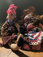 Two dolls. On the left, cannot make up zir mind about pronouns, wears a wonderful red feather headpiece. On the right, She / her in a lovely brown and white crocheted dress and enormous Pippi LongStocking Braids