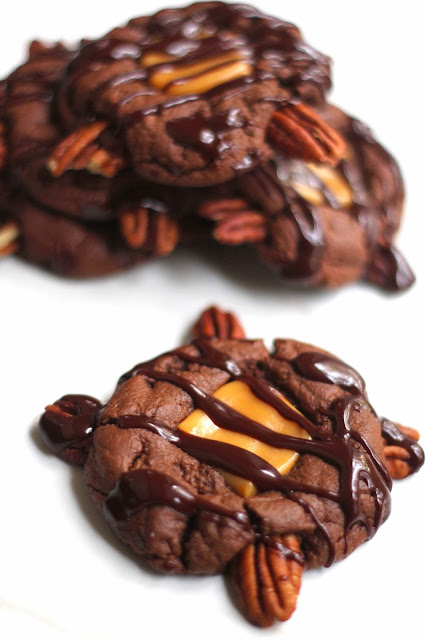 A turtle cookie in the foreground drizzled with chocolate sauce on top and a stack of turtle cookies in the background