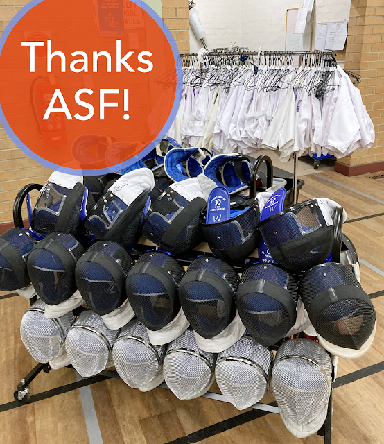 Photo with rows of fencing masks on a low rolling rack in the foreground, behind which is a higher rolling rack with a large number of fencing plastrons hung on coathangers. In the top left of the image is a large orange dot with a pale purple border with the text 'Thanks ASF!' in white on the orange dot.