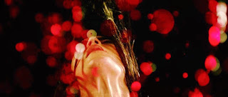 Em close de rosto num fundo preto, uma mulher branca joga a cabeça para trás, fazendo voar o cabelo liso e preto. O que parecem ser diversas gotas vermelhas brilham espalhadas pela imagem.