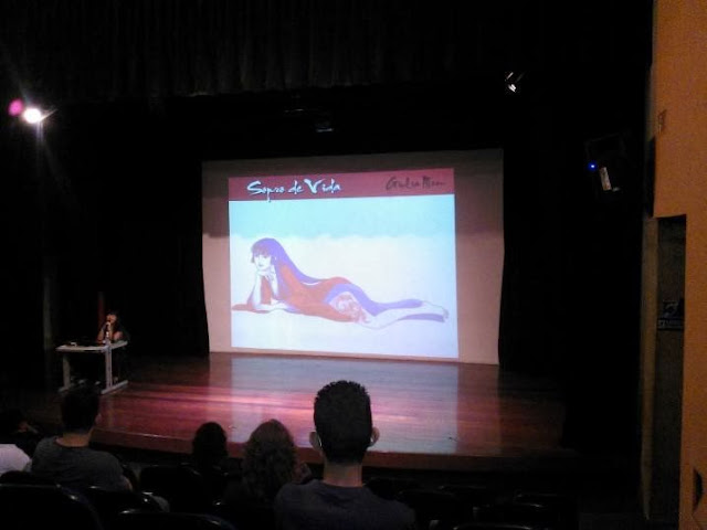 Foto do auditório da Estácio Uniradial com um slide de Kaori projetado na parede, referente a palestra de Giulia Moon, ao centro do palco