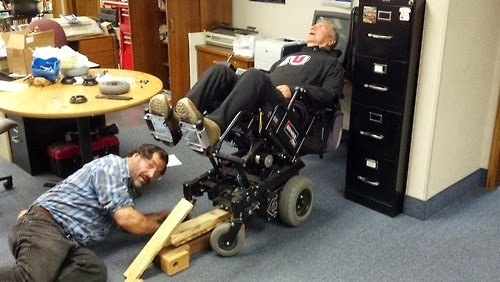 Man in reclined electric wheelchair with another man repairing the chair