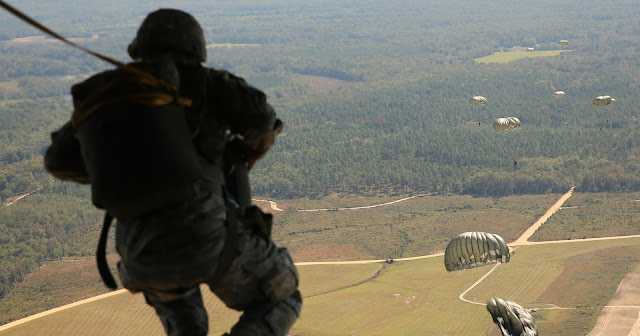 A service member in uniform prepares to jump out of an air vehicle. There are other service members with parachutes flying through the air.