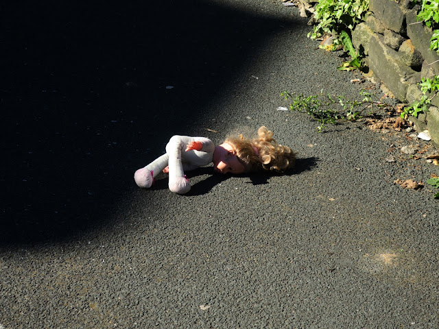 Cloth doll abandoned on pavement between shadow of house and dry stone wall with plants.