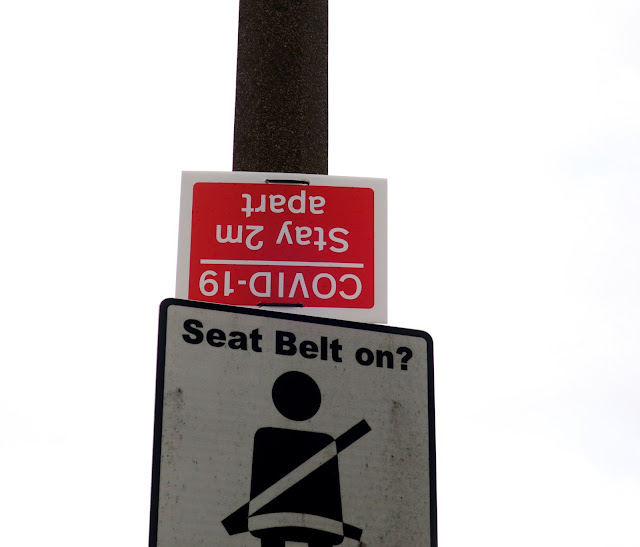 Covid 19 distancing notice put upside down on a post above a use-your-seat belt notice.