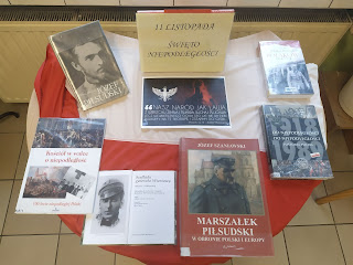 Na stoliku leżą książki o Piłsudskim. Po lewej stronie flaga Polski. Na środku: 11 listopada Święto Niepodległości.