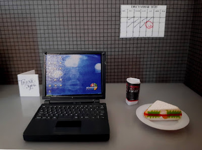 Close up of a one twelfth scale modern miniature office cubicle with a laptop, a sandwich and takeaway coffee, a thank you card and a December 2020 calendar with 11 December circled with a red smiley face.