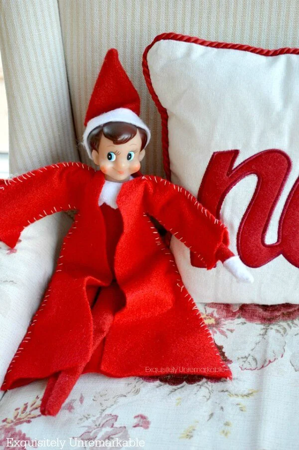 Elf on the shelf with a DIY coat sitting on chair with Christmas pillow