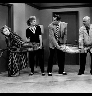 Lucy and Ethel carrying a large fish and staring surprised at Ricky and Fred who are also carrying a large fish.