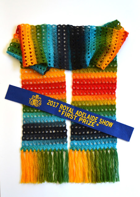 The broomstick lace rainbow scarf is laid flat against a white background with the fringed ends pointing down. the centre of the scarf is draped at right angles to the ends. A blue ribbon is laid diagonally across the scarf. The writing on the ribbon is in yellow "2017 Royal Adelaide Show *First Prize*" with the Royal Agricultural Society's coat of arms.
