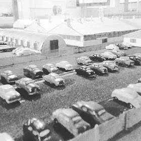 Model of a factory. In the foreground are lines of cars parked outside it.