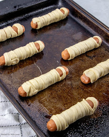 Hot dog wrapped in crescent roll strips to make a mummy lined up on baking sheet