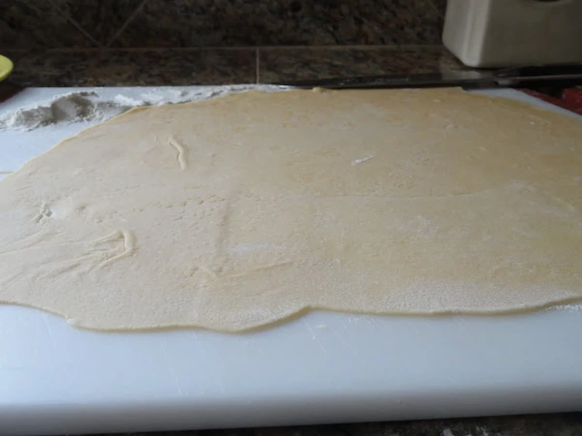 Rolled out Homemade Egg Noodle Dough on a white cutting board with a small pile of flour in the back ground.