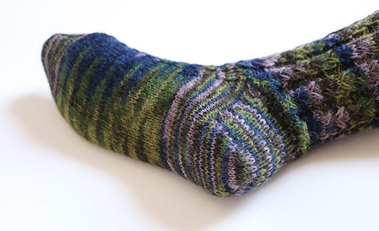Bottom view of foot wearing hand knit wool sock in blues, greens and purples on a white background.