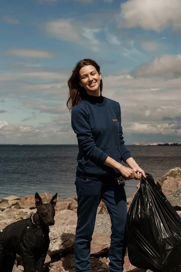 Woman smiling while holding a trash bag during a coastal cleanup, with a dog beside her on a rocky shore.