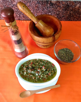 Un tazón que contiene, salsa chimichurri, junto a un envase con orégano seco en hojas, un mortero de madera, y una botella decorativa, alta y delgada, que contiene diferentes tipos de condimentos.