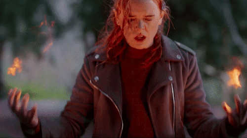 Gif: a personagem Bloom, uma garota ruiva em uma roupa vermelha com um casaco marrom-avermelhado, erguendo os braços e fazendo chamas surgirem de suas mãos e no fundo uma floresta.