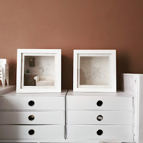 Two sets of white cardboard storage drawers against a mushroom-brown wall. On top are two white box frames, one containing a 1/12 scale sofa, side table, art work and pot of catci, the other is empty. Next to the drawers is a white ring binder.