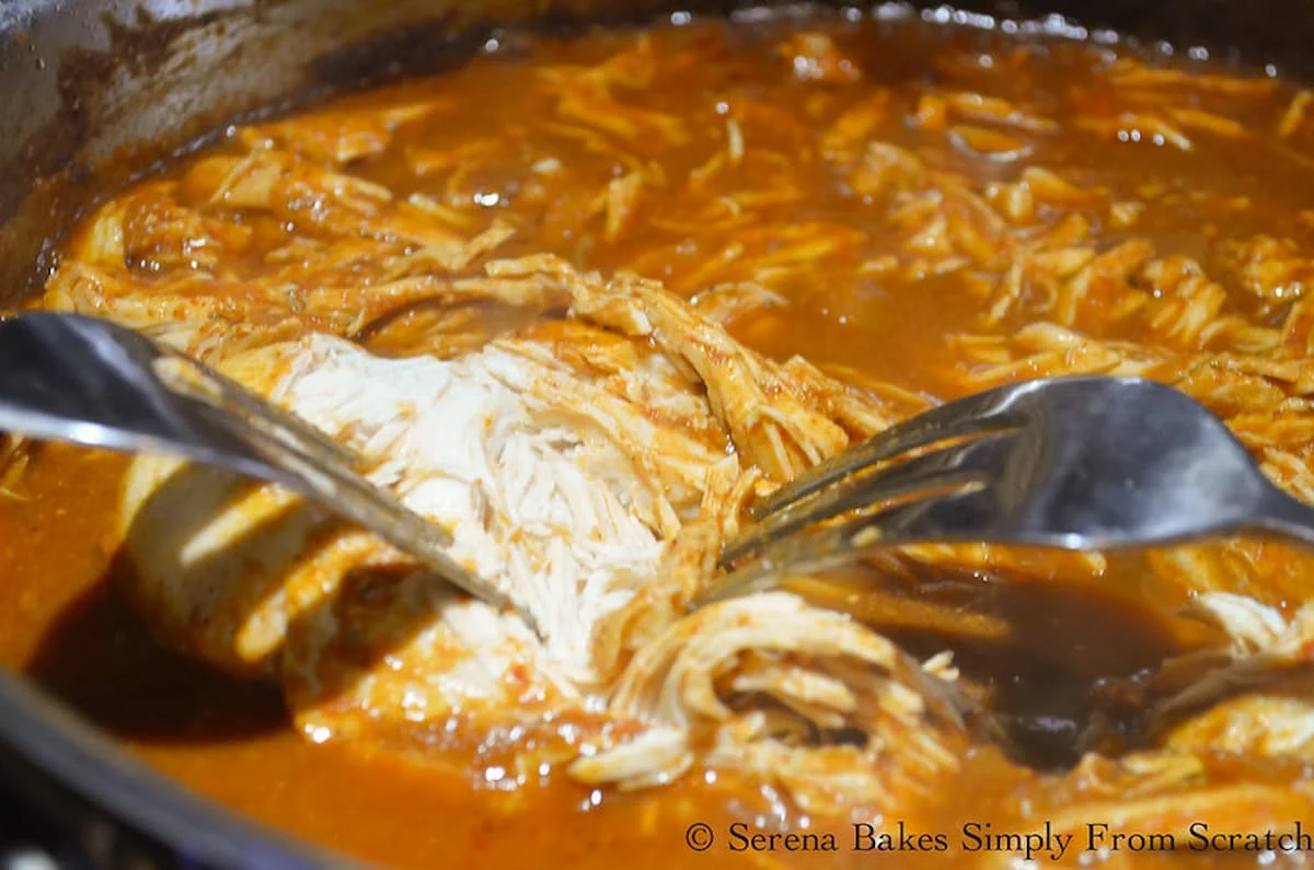 Chicken being shredded with two forks in Red Enchilada Sauce in a stainless steel pan.