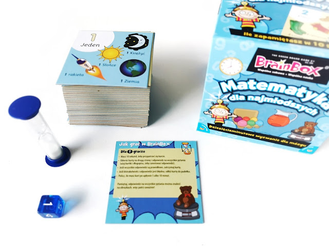 na zdjęciu pudelko gry brain box matematyka dla najmłodszych, obok ley stos kartonowych kart z pytaniami, klepsydra oraz niebieska kostka sześciościenna