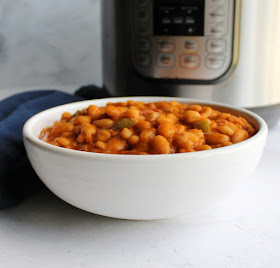 Baked beans are a classic side dish for good reason. This recipe flavors delicious baked beans with maple and bacon. They start with dried beans and can still be made in a couple of hours with the help of a pressure cooker. looking across a bowl of homemade maple baked beans with pressure cooker in background