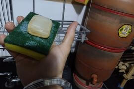 Imagem de alguém segurando uma bucha com a mão direita, ao fundo uma pia negra e azuleijos brancos de cozinha, presença de um sabão em barra sobre a Bucha de marca Scotch Brite