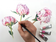A hand holding a paintbrush painting pink and white flowers. There are also tubes of paint on the lower right.
