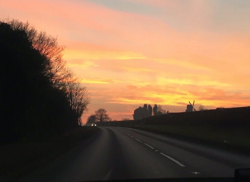 Morgan's Milieu | Leaving the Novel Behind: A photograph of the sky at sunset. A darkened windmill in the distance and a car on the road.