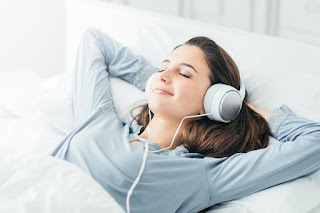 Caucasian woman with brown long hair and a blue t-shirt she is laid in bed with her hands behind her head her eyes are closed and  she is wearing white headphones