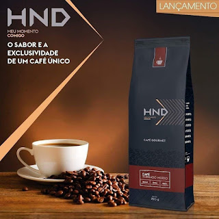 Imagem da embalagem do café gourmet Hinode ao lado de uma saborosa xícara de café entre os grãos de café Hinode. 