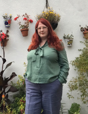 A red-headed woman looking into the camera. She has her hands in her trouser pockets and is wearing a green blouse.